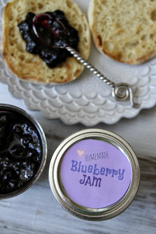 overhead shot of blueberry jam in a jar with the lid displayed too, The lid has a purple label. english muffin with spoon of jam on it on a plate.